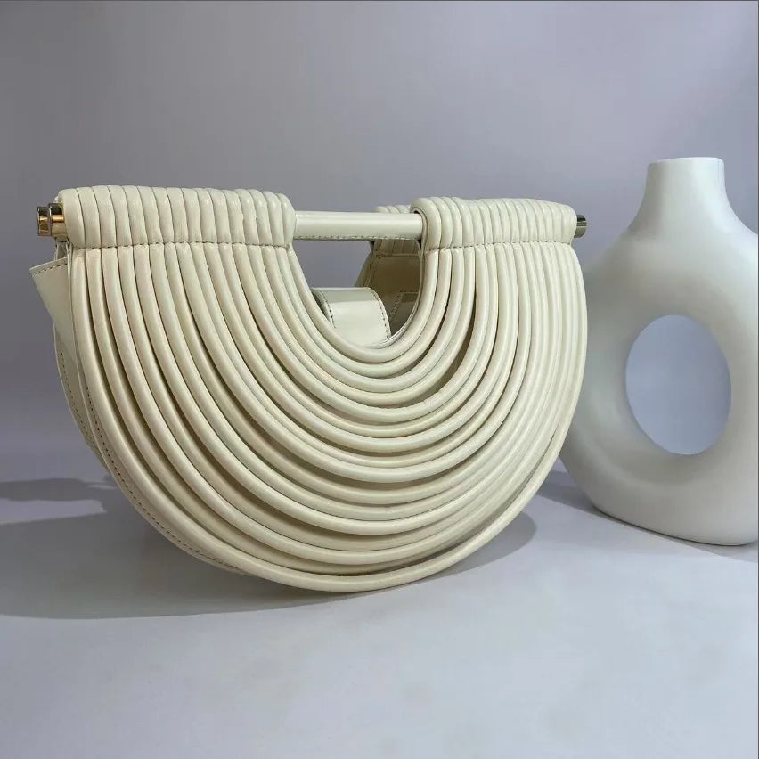 Cream-colored handbag with braided design and gold accents next to a white vase on a light gray background